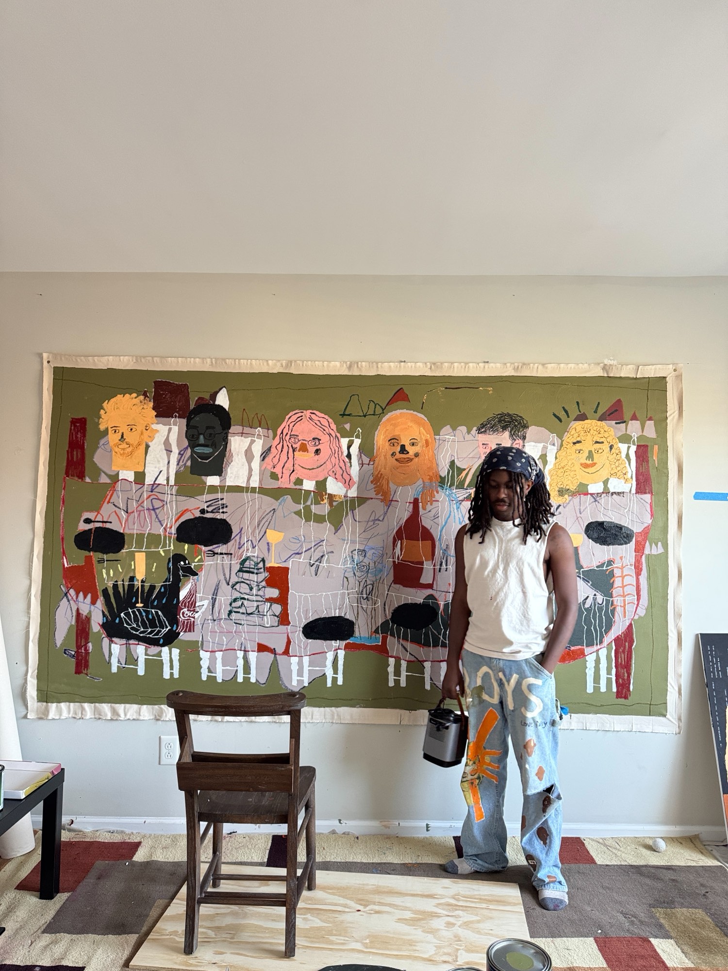 Bhare in front of a wide unstretched canvas of faces, holding a paint can